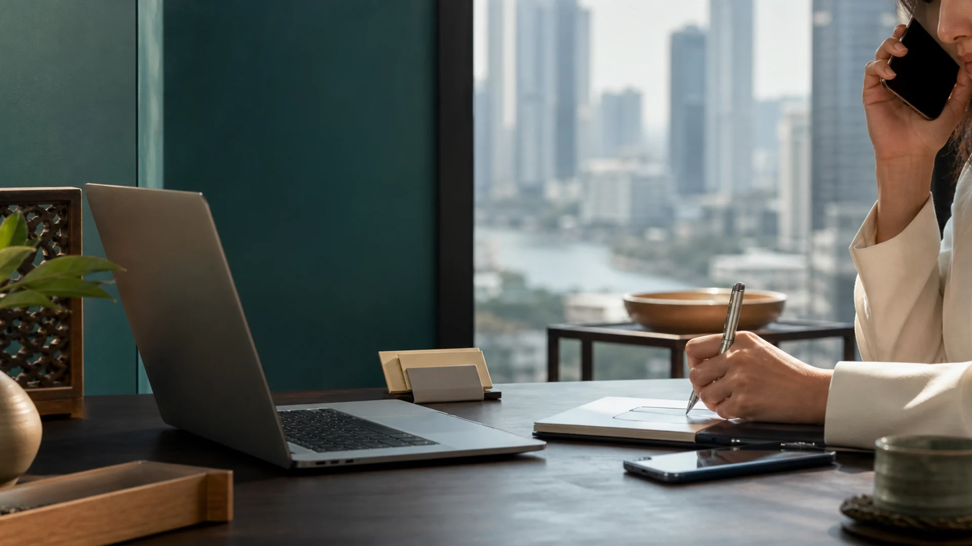 A laptop and phone on a wooden desk with the Bangkok skyline visible through a window at sunset